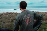 A person viewed from behind wearing a long-sleeved green t-shirt with a circular emblem design, standing in an outdoor setting with grass and a body of water in the background.